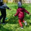 A child dressed in a red jacket and trousers running after a ball on green grass