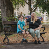 A foster carer and a child are sat on a bench in a park laughing together.