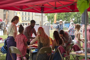 A group of people doing an activity while sitting underneath a red gazebo on College Green. Other stalls can be seen in the background.