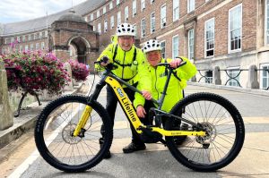Two people dressed in high vis stand behind an electric bike outside City Hall. 
