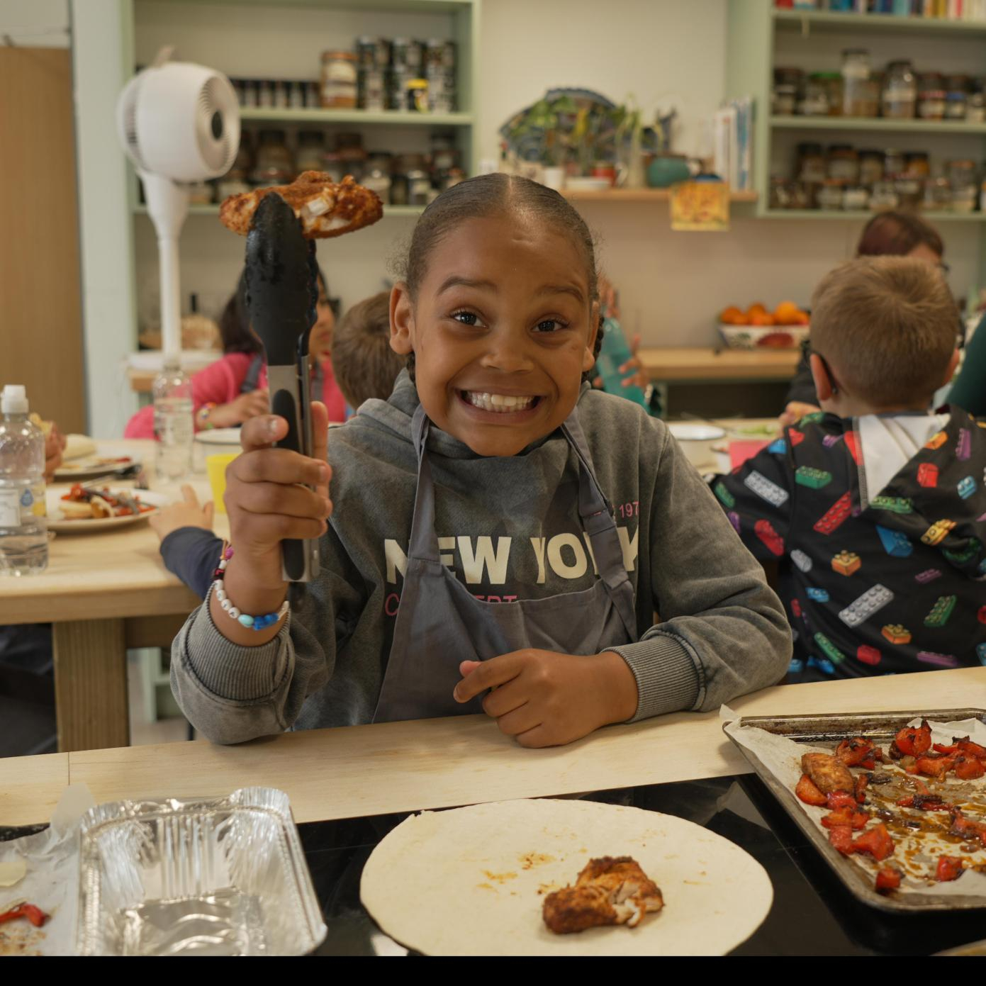 A child in a kitchen is holding food with tongs, looking straight ahead and smiling. Other people in the cooking session are in the background.