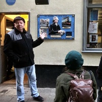 A walking tour guide stands in historic square pointing. People stand and listen.
