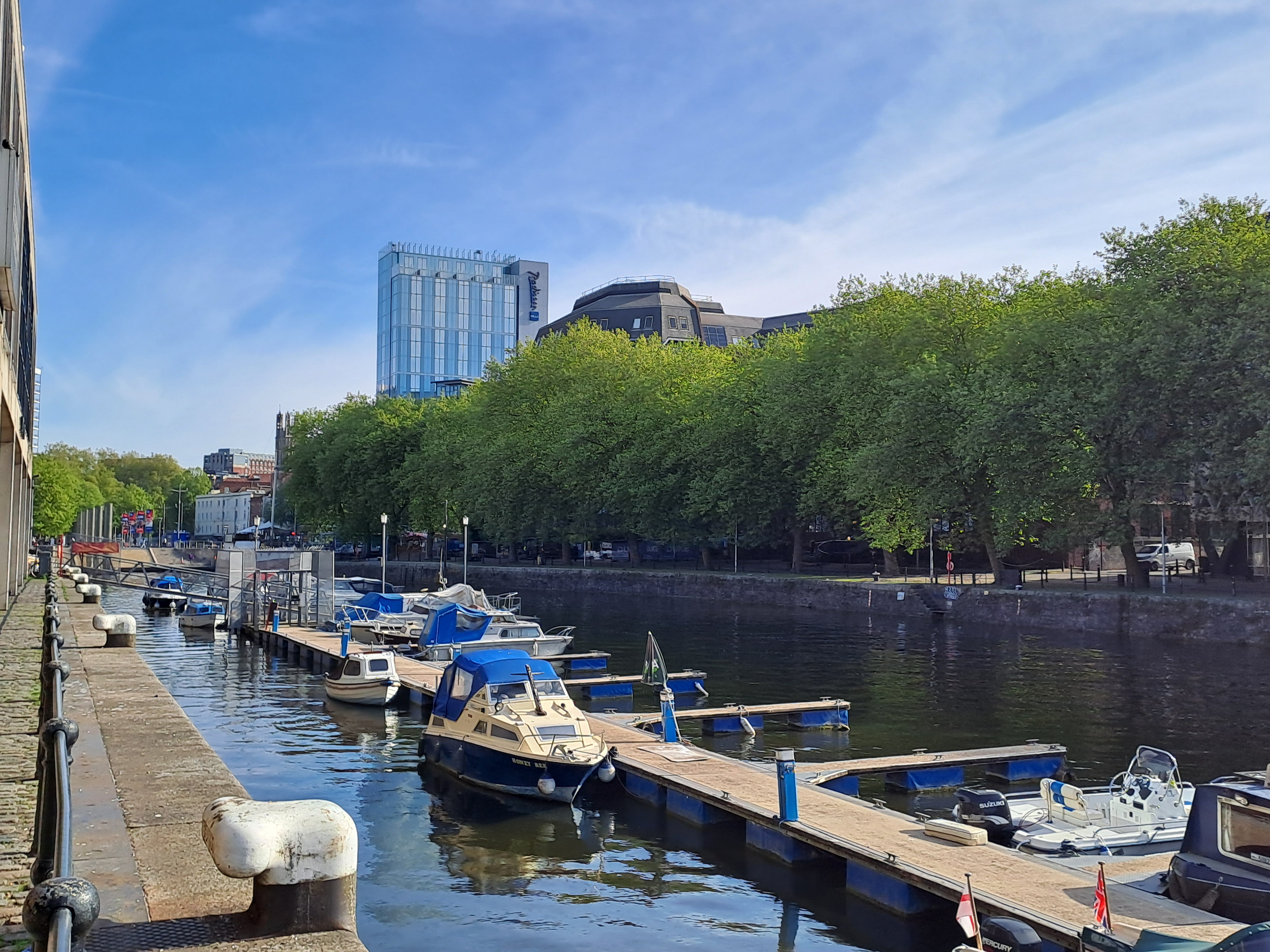 Some small boats moored in the harbour, trees and a big blue glass building in the back