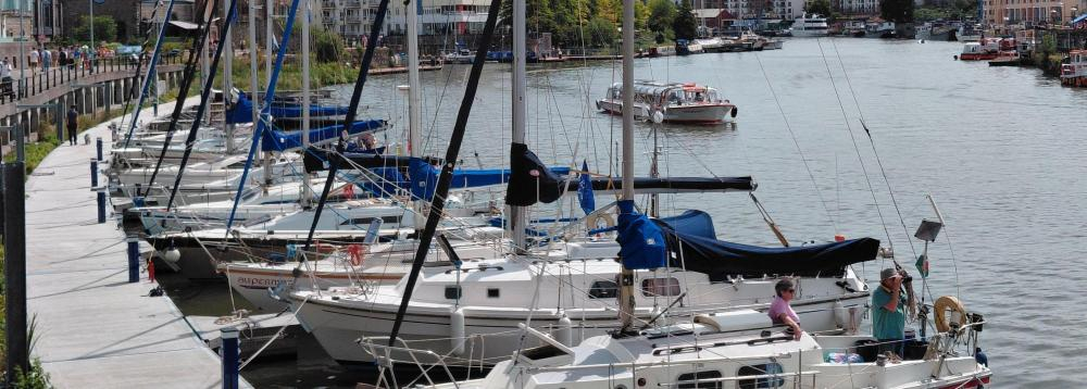 Some boats in mooring platforms on the harbour, water in the background and some buildings