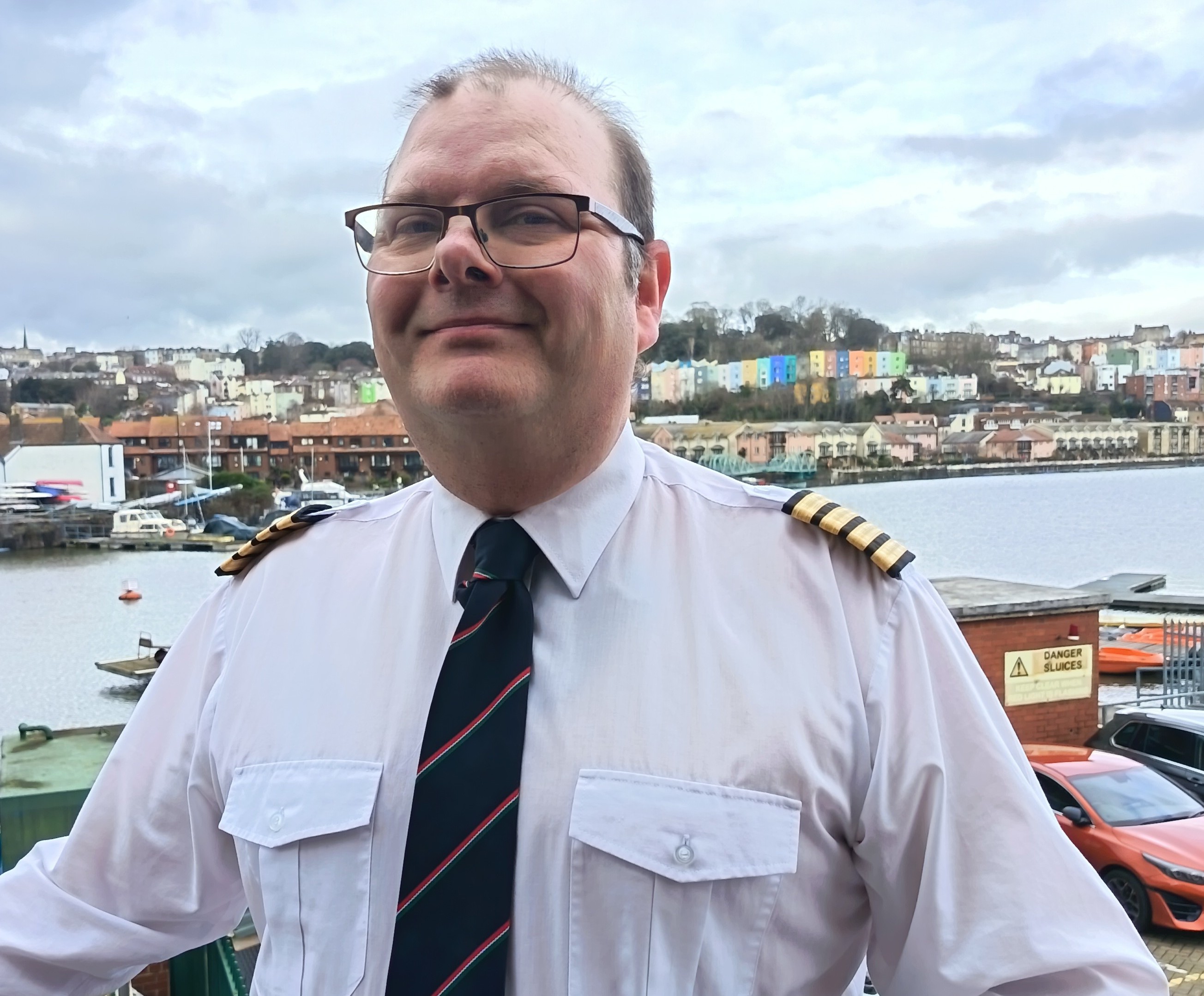 A smiling man wearing glasses and a white shirt. In the background, the harbour and colourful houses
