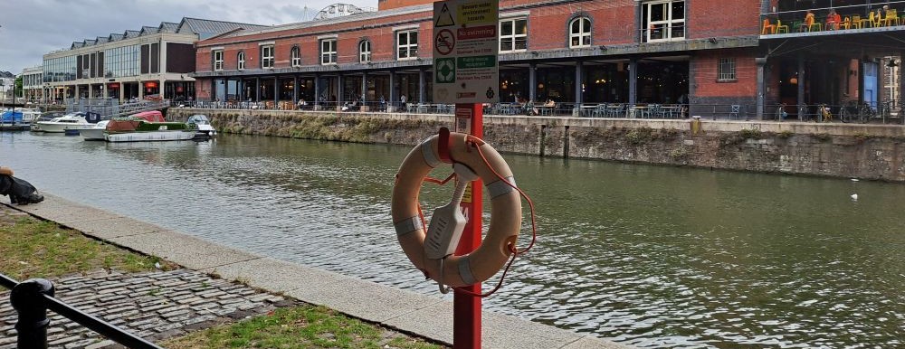 A life saving ring tied to a red post. The water, boats and bars in the background