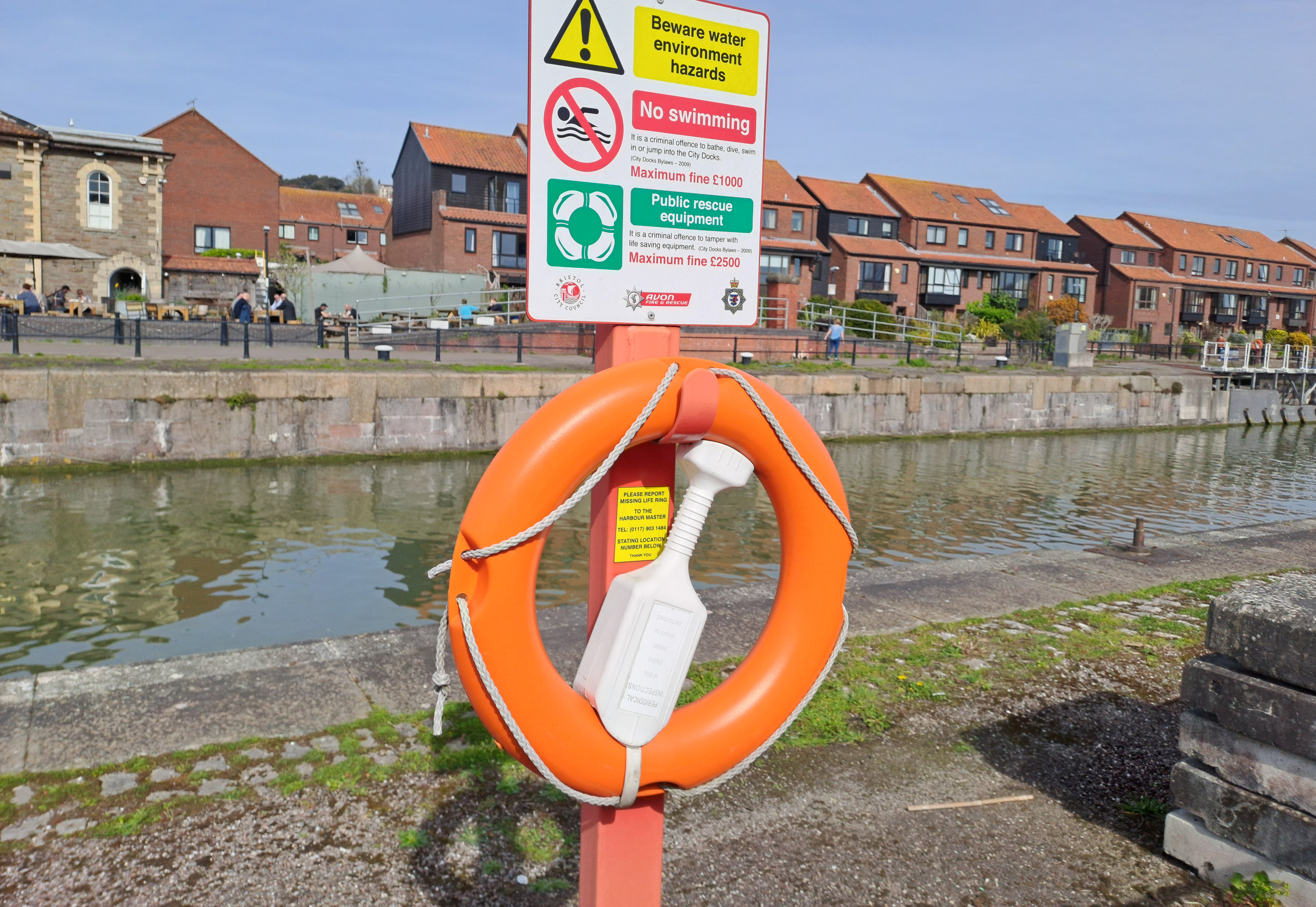 An orange life saving ring tied to a red post. The water, boats and bars in the background