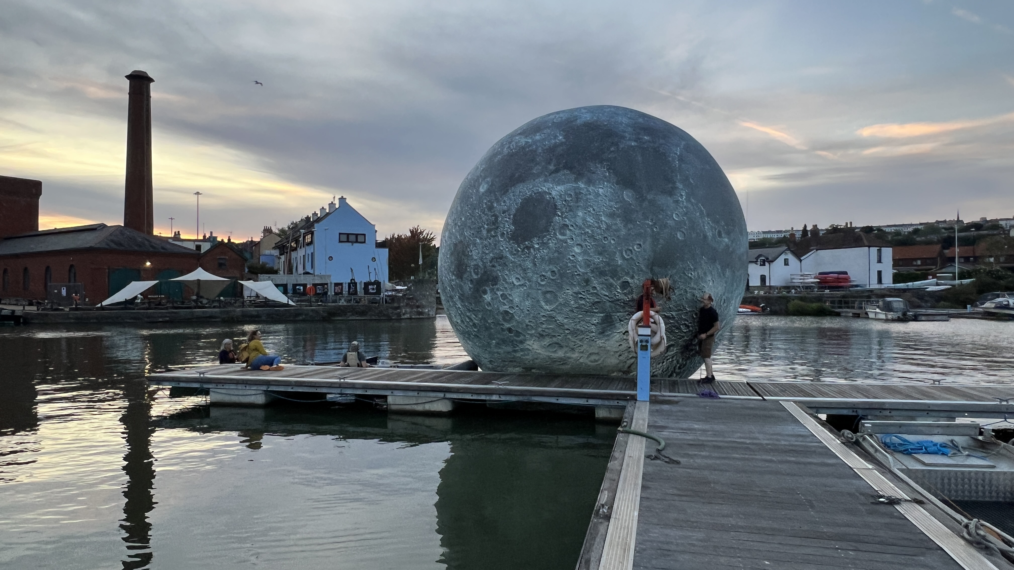 A large moon shape at the edge of a slipway, the harbour water in the background and some white buildings off shore