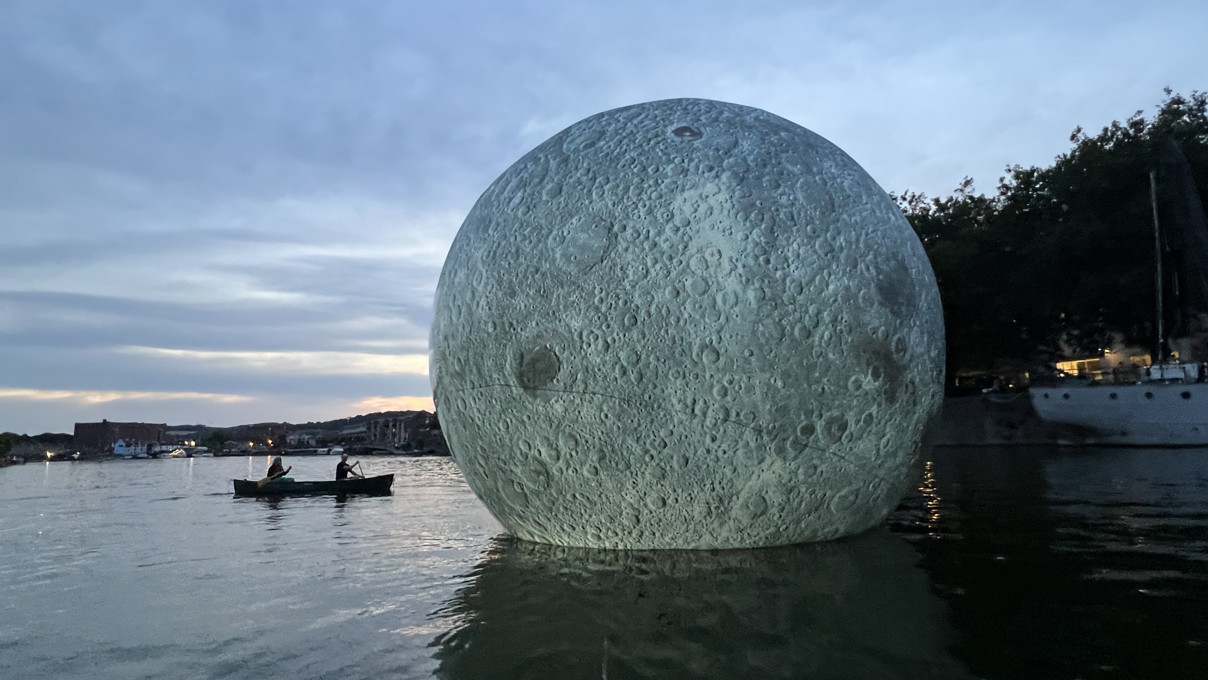 A large moon shape on the water at dusk, a boat with two people next to it.