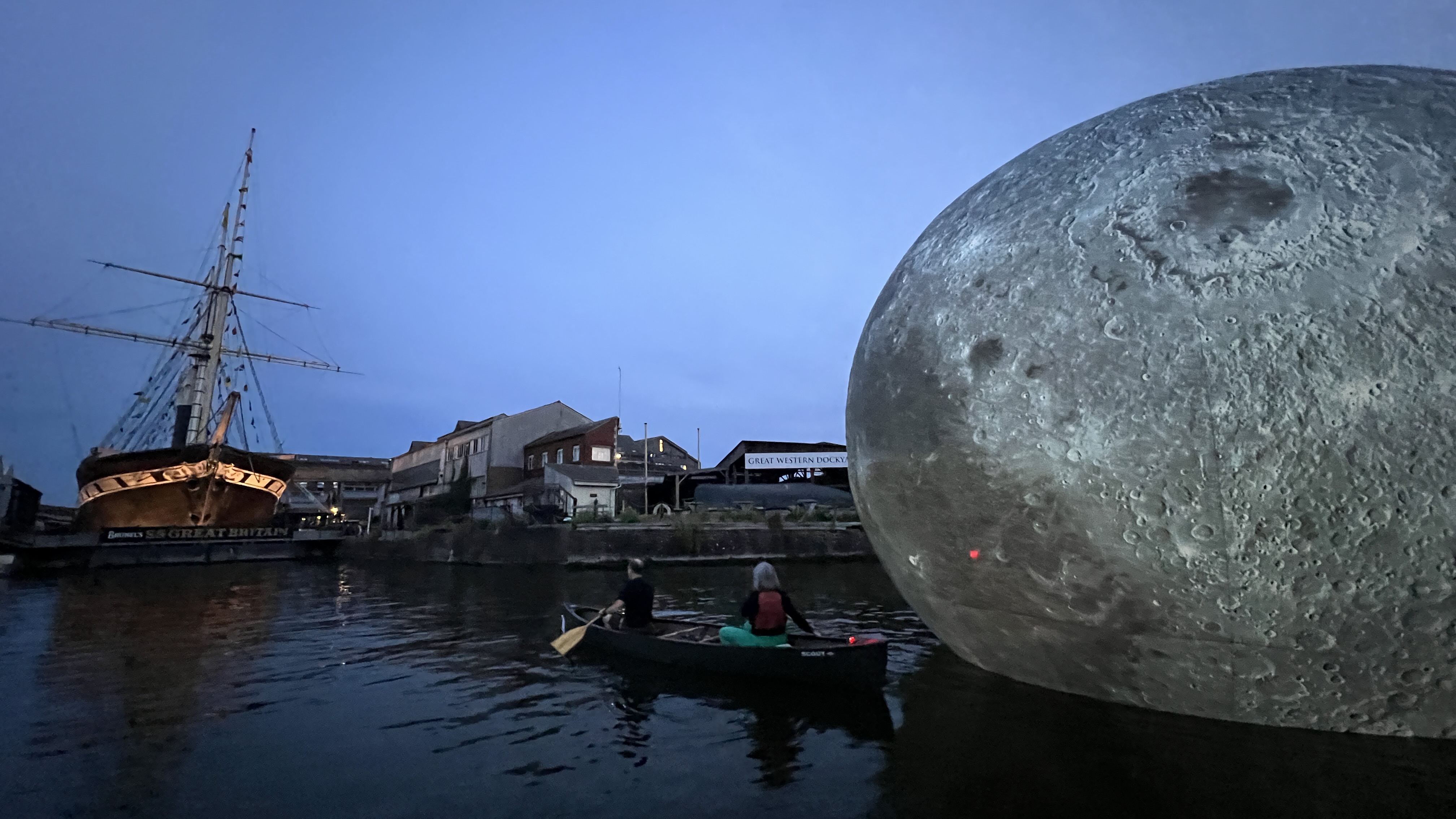 A large moon shape lit up on the water, a small boat to the side and a big brown boat with masts in the background