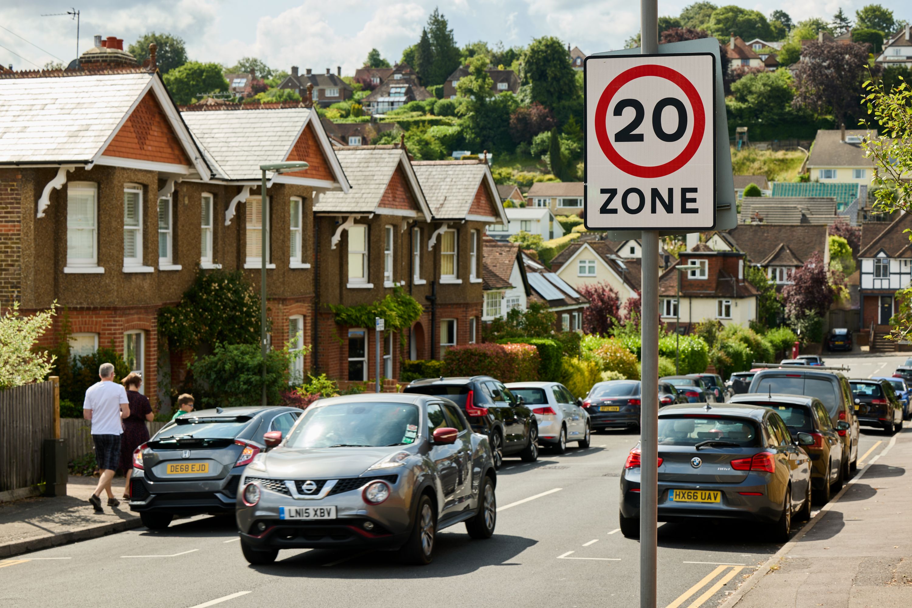 20 is plenty sign in a residential area, cars on the road, houses and people walking on the pavement