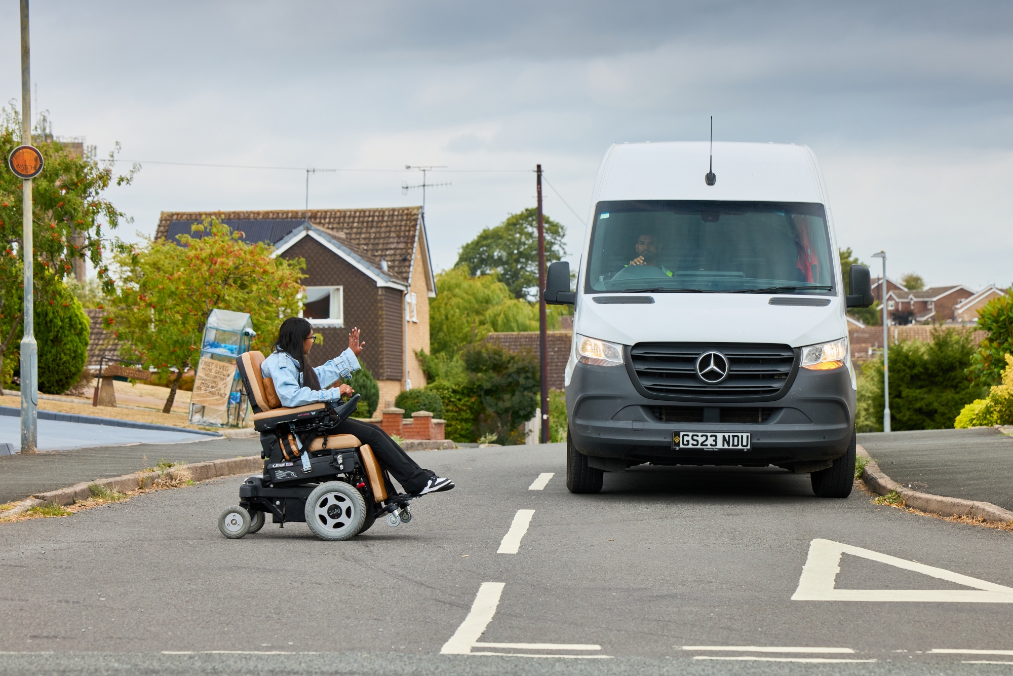 A young girl in a wheelchair crossing a road, waving to a white van coming toward her
