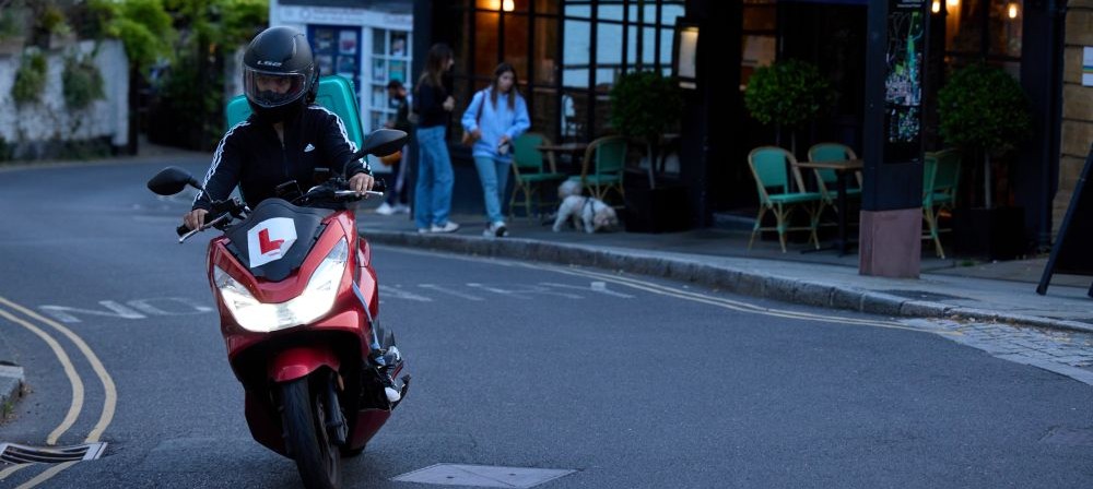 A person on a moped with a learner plate on the front going down the road, people walking down the road behind them and a cute little dog