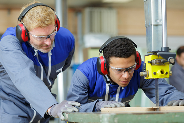 One man supervises another while he uses a table saw in a factory
