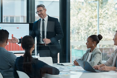 A group of people in a meeting with someone presenting some stats in a graph.
