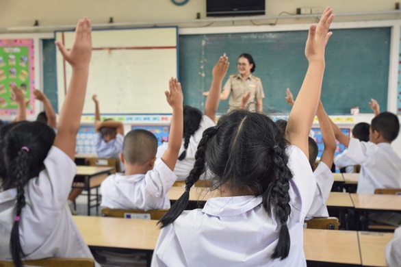 A classroom full of children raising their hands to answer a question from the teacher.