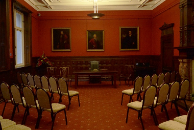 A room with wood panelling, period paintings, a large fireplace and two sets of three rows of chairs